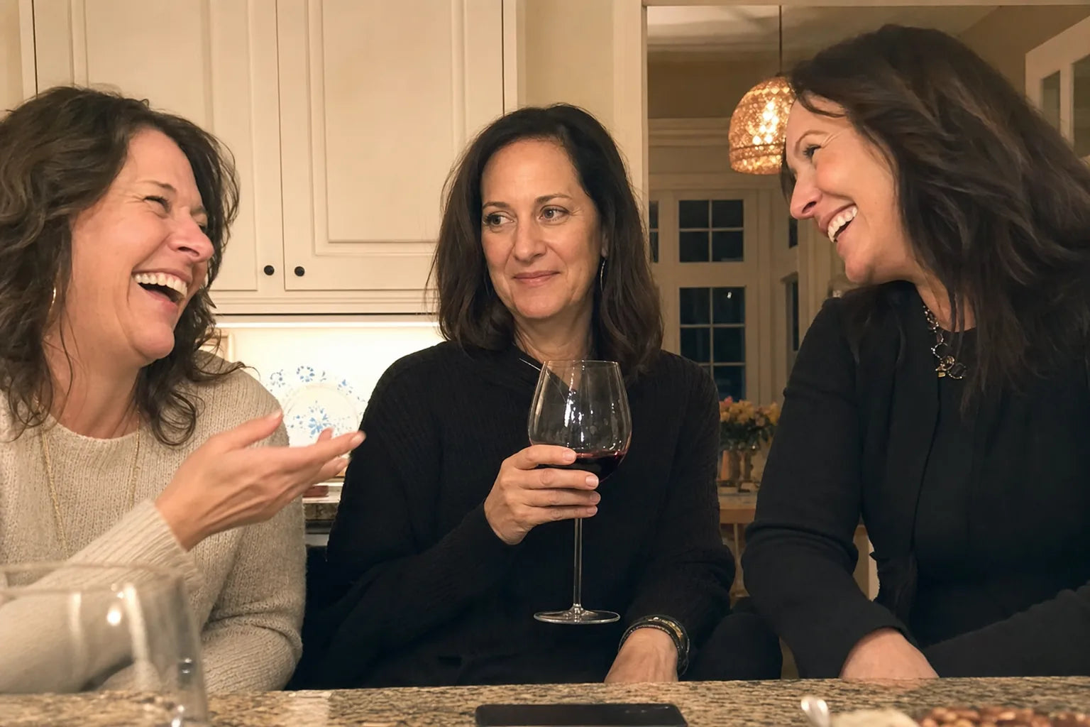 Three women at a kitchen island on a wine night, two laughing and one with a polite smile that does not reach her eyes