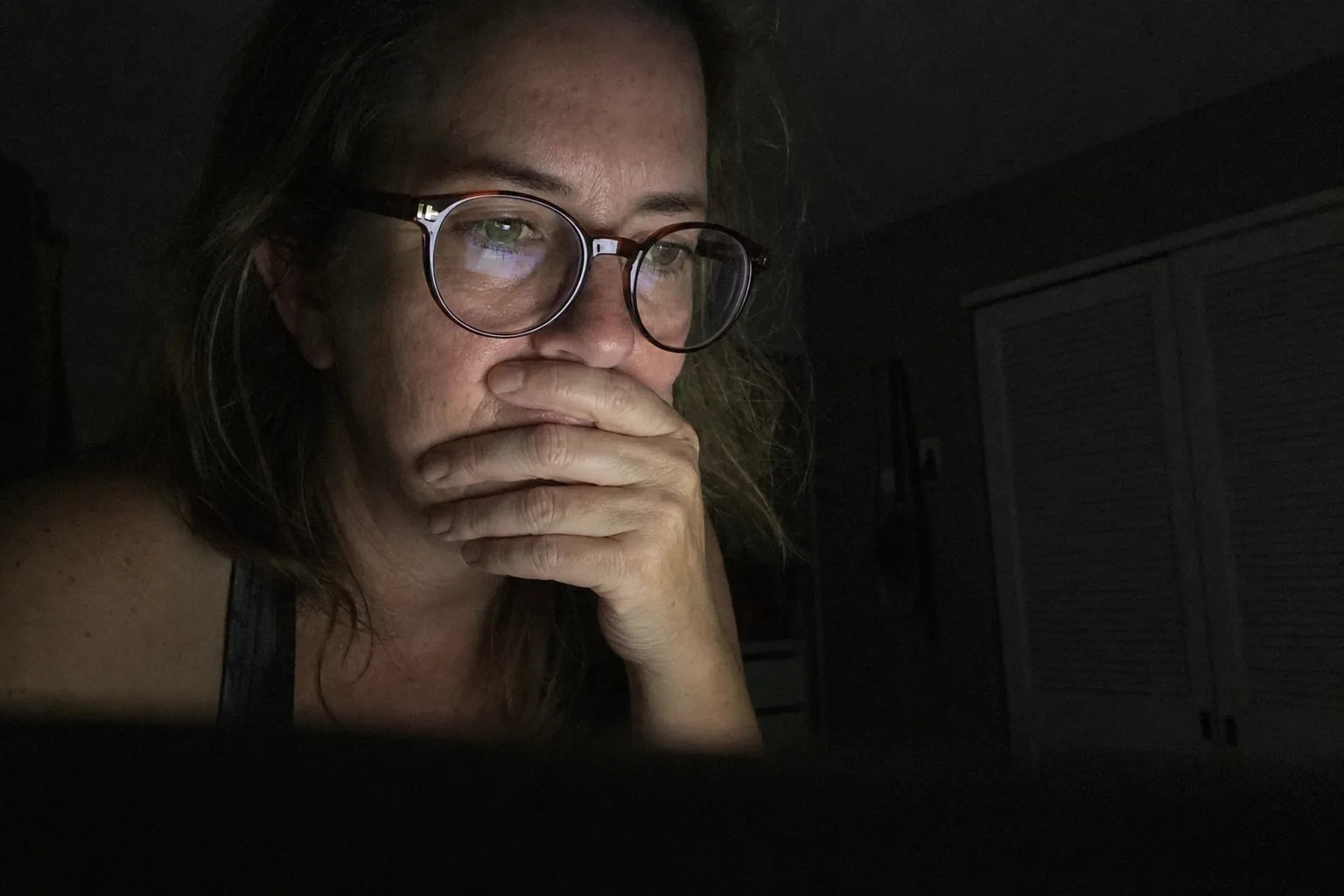 Woman at 11pm lit only by laptop glow, one hand over her mouth, a forum page faintly reflected in her reading glasses