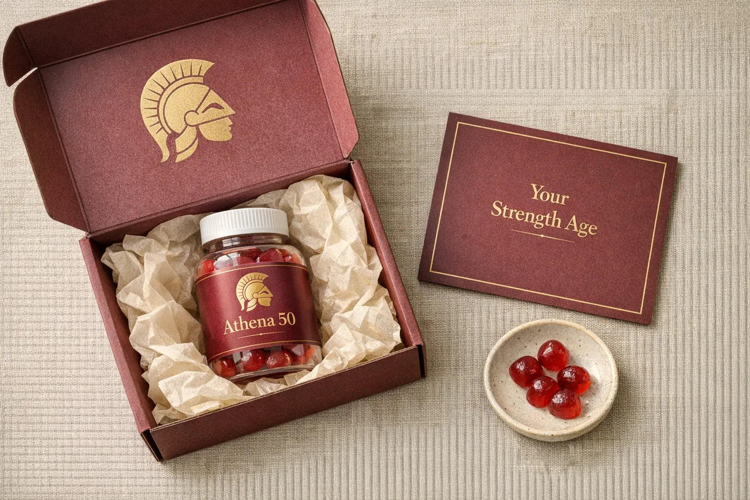 Athena 50 bottle beside the Strength Age Card and two ruby gummies on marble with a gold 90-Day Stronger Guarantee seal